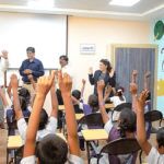 Classroom with pupils hands raised. Teachers seen smiling at the front. 