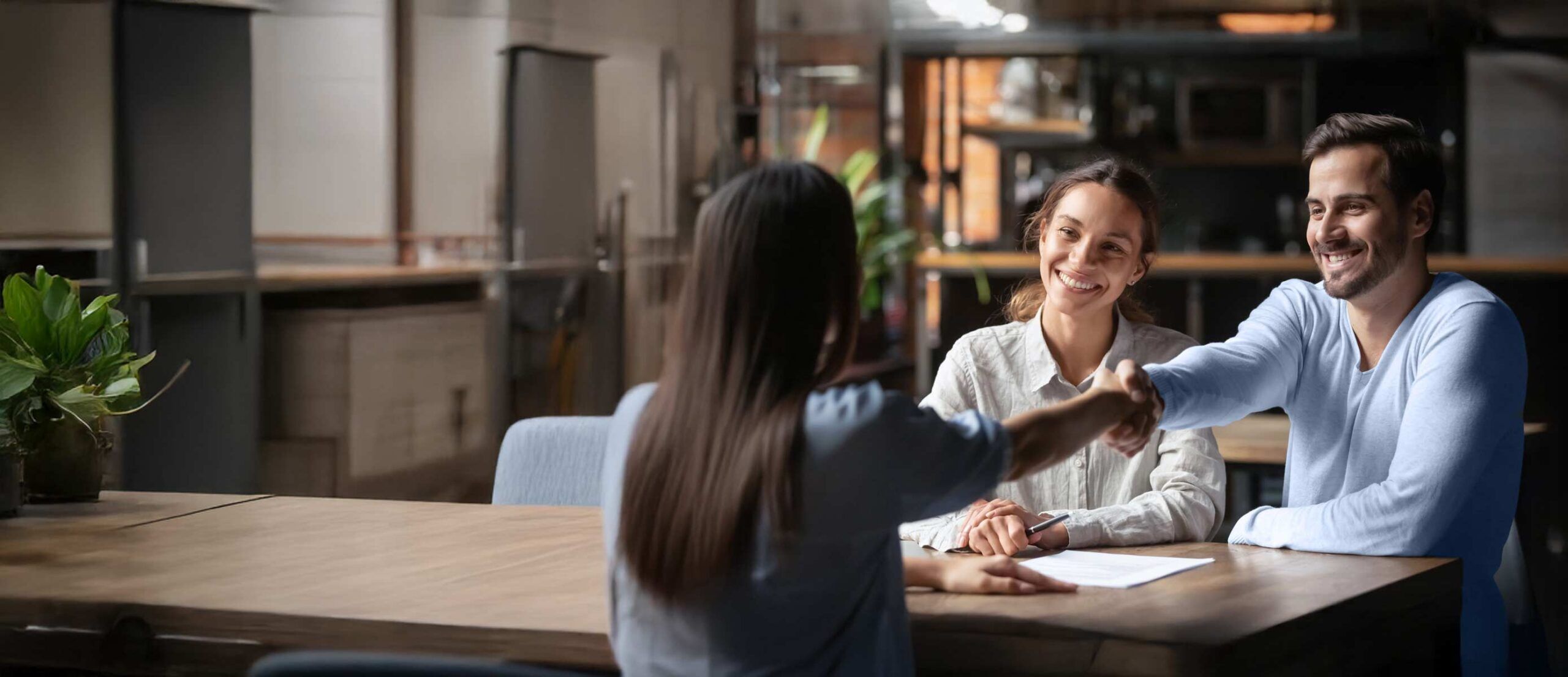 A person seen from behind is shaking hands with a man. Next to the man is a woman. They are smiling and sitting at a long table.